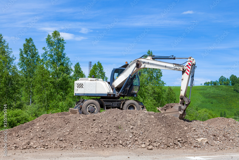 A working white escalator with a blue cabin with a lowered bucket raking on a large pile of land against a background of green trees and sky during the construction of a suburban highway. A working white escalator with a blue cabin with a lowered bucket raking on a large pile of land against a background of green trees and sky during the construction of a suburban highway.