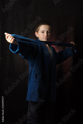 A boy stands on a dark background in a blue kimono and shows his belt. Martial arts.