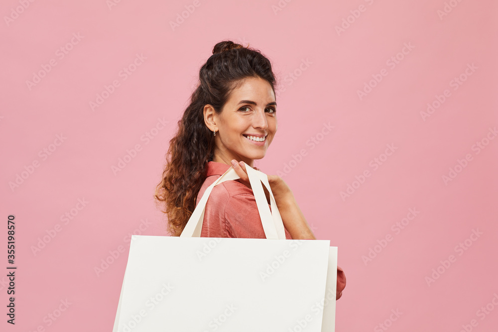 Portrait of happy young woman smiling at camera and holding white paper bag isolated on pink background Portrait of happy young woman smiling at camera and holding white paper bag isolated on pink background