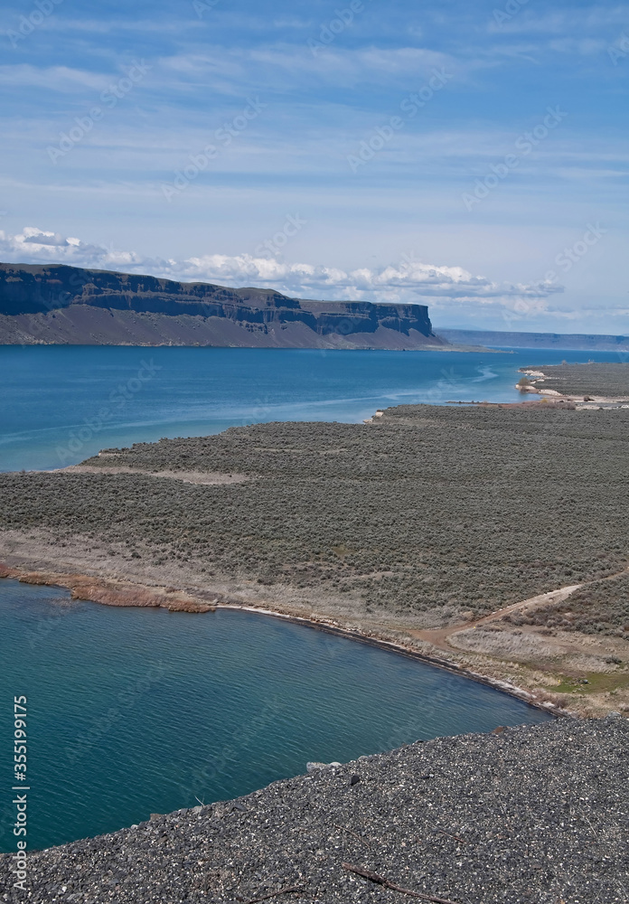 Desert lake landscape shows high mesa bluffs in the distance, and cool, turquoise colored water in the lakes beneath.  Vertical image of Banks Lake, in Grant County, Washington, America.