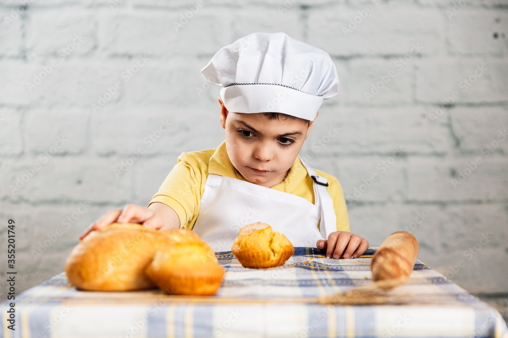 un niño vestido con gorro y delantal de panadero toca el pan con curiosidaden la sesion de fotos con el pan y las magdalenas. escena de aire rustico. estilo de vida, panadería. un niño vestido con gorro y delantal de panadero toca el pan con curiosidaden la sesion de fotos con el pan y las magdalenas. escena de aire rustico. estilo de vida, panadería.