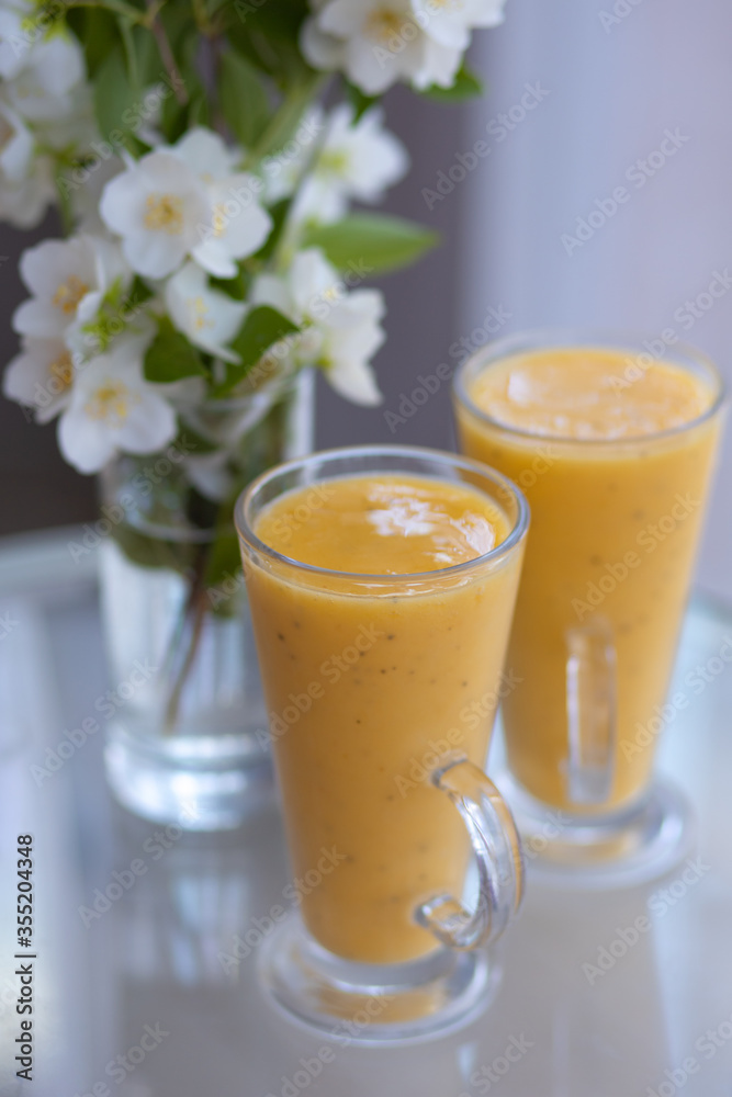 two yellow smoothie cocktails in high glasses on a glass breakfast table in the morning with white flowers. Side view. Summer Spring two yellow smoothie cocktails in high glasses on a glass breakfast table in the morning with white flowers. Side view. Summer Spring