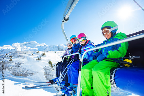 Low view portrait of four happy kids in vivid ski outfit lifting on chairlift on the mountain together and smiling