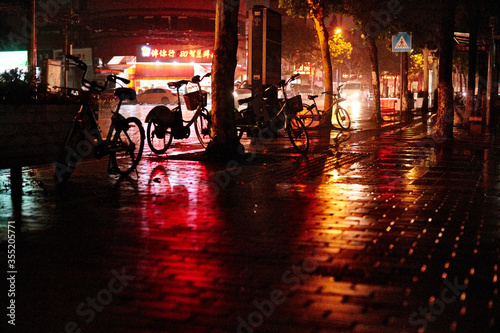 Bicycles in the rain on the night streets of Shanghai, China. Lots of colours reflecting on the ground in this grainy nighttime photo. Dark and moody © Chris