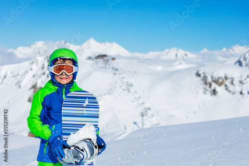 Close portrait of a boy in sport helmet hold snowboard glasses over bright sunny mountain snow tops on background
