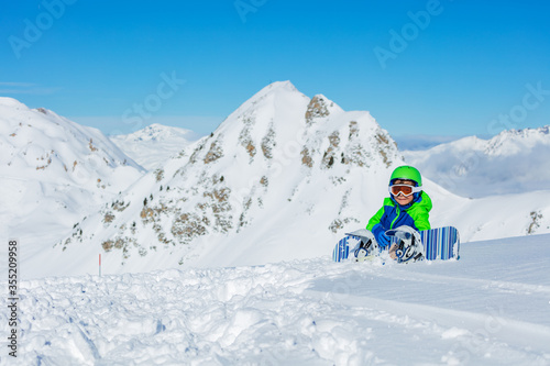 Portrait of a little snowboarder sit in snow with snowboard over mountain in bright blue and green sport outfit mask, helmet