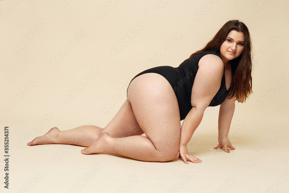 Plus Size Model. Full-Figured Woman In Black Bodysuit Portrait. Crawling Brunette Posing On Beige Background And Looking At Camera. Body Positive Concept.