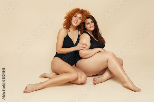 Diversity. Models With Different Figure And Size Portrait. Female Friends Sitting On Floor. Smiling Brunette And Redhead In Black Bodysuits Posing On Beige Background. photo