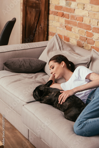 smiling woman lying on sofa and touching black french bulldog