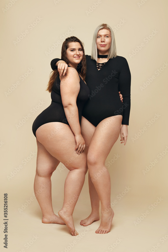 Plus Size Models. Full-figured Women Full-Length Portrait. Brunette And  Blonde In Black Bodysuits Posing On Beige Background. Body Positive  Concept. Stock Photo