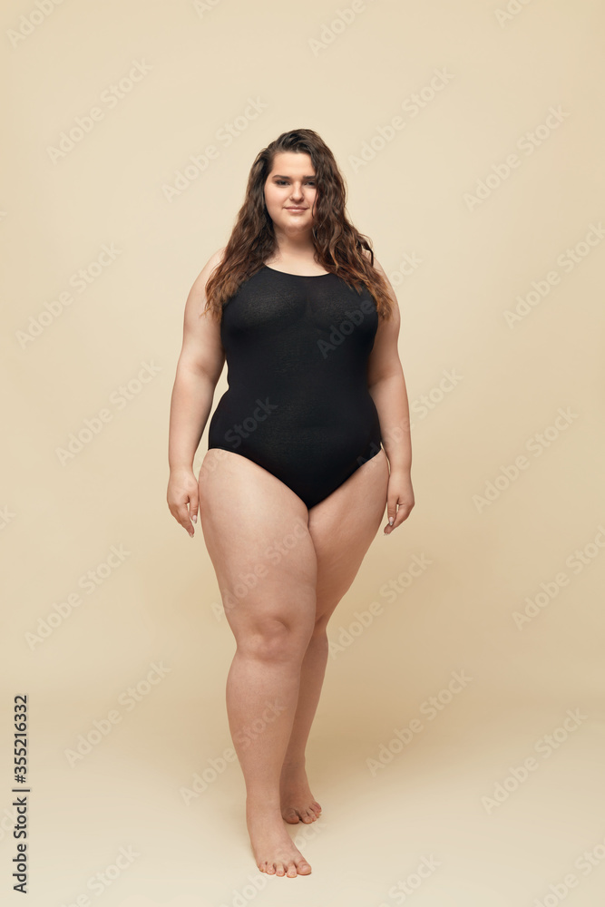 Plus Size Model. Big Woman In Black Bodysuit Full-Length Portrait. Brunette  Standing And Looking At Camera. Body Positive Concept On Beige Background.  Stock Photo