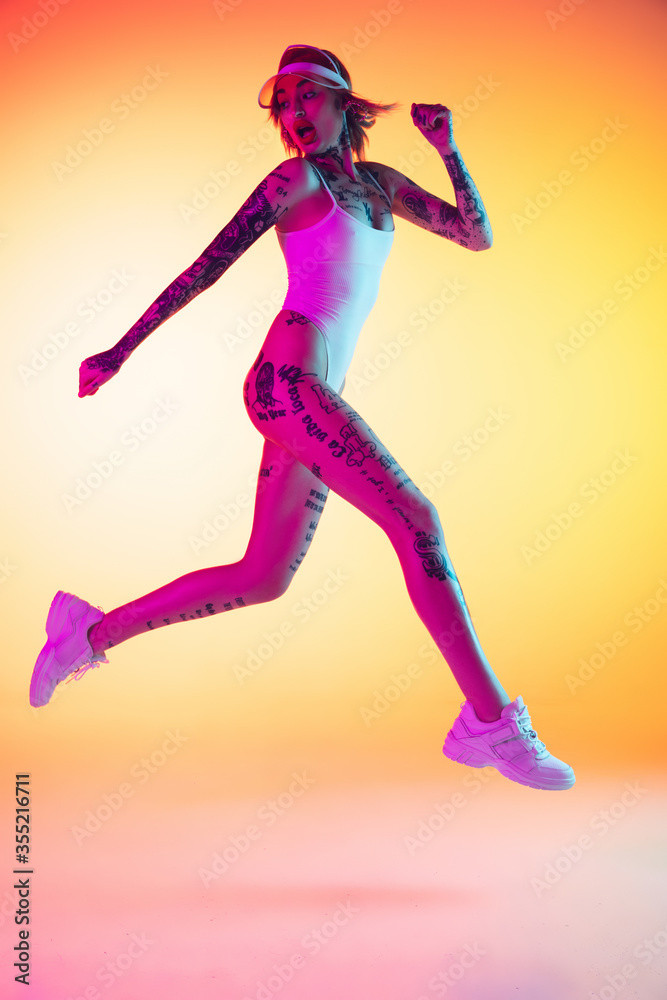 Young caucasian woman in swimsuit and beach hat jumping, running on gradient yellow background in neon. Beautiful model with tattoos. Human emotions, sales, ad concept. Resort and vacation, summertime