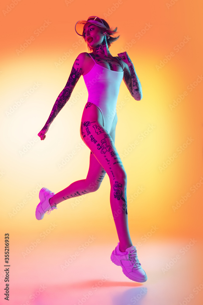 Young caucasian woman in swimsuit and beach hat jumping, running on gradient yellow background in neon. Beautiful model with tattoos. Human emotions, sales, ad concept. Resort and vacation, summertime