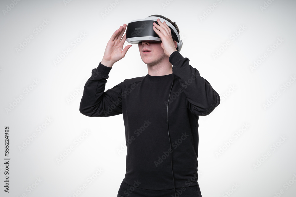 Young man in black shirt uses VR glasses in white studio, dreamily looking away and touches glasses with hands, waist-height portrait Young man in black shirt uses VR glasses in white studio, dreamily looking away and touches glasses with hands, waist-height portrait