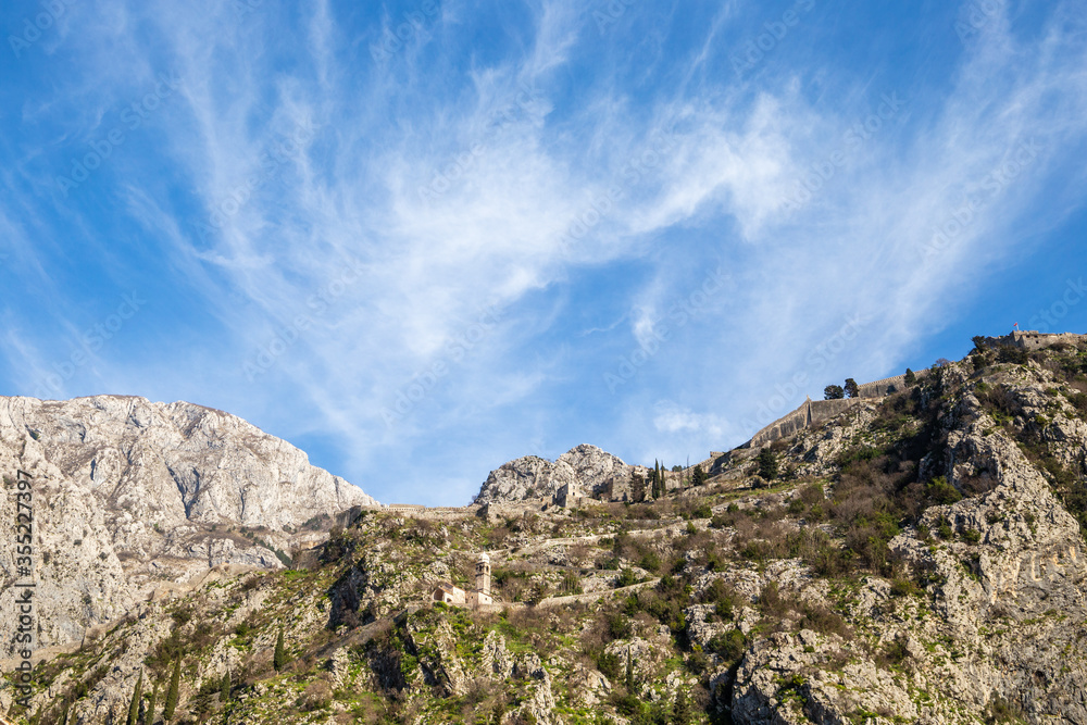 Mountain ridge featuring a medieval fortification wall under blue sky with high cloud formations. Rough and rocky terrain in the Mediterranean. Hills overlooking Kotor Bay in Montenegro.