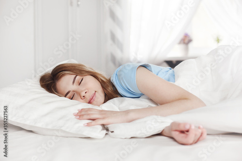 A beautiful young girl in blue pajamas is sleeping on a bed in her bright bedroom at home in the morning.