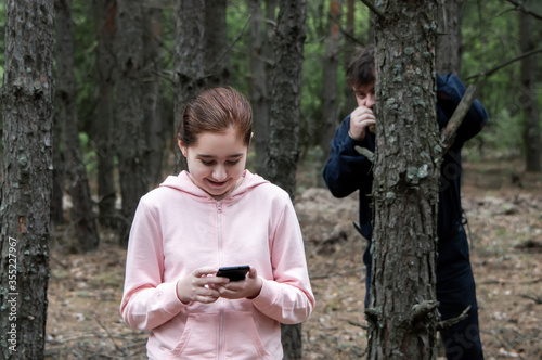 Foto A maniac watches from behind a tree in the forest for a teenage girl in a tracksuit with a mobile phone in her hands