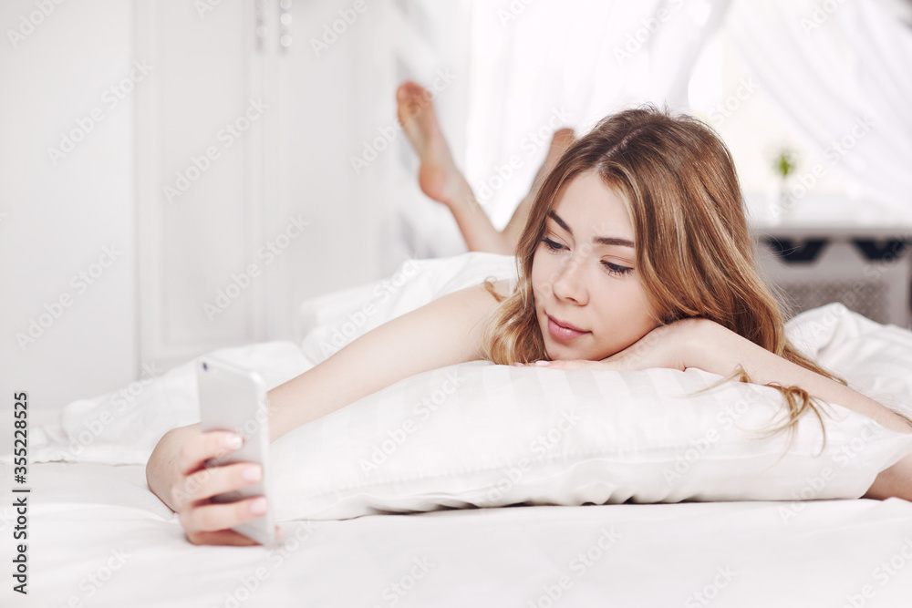 Cute smiling girl looks at the phone, lying on the bed. A girl reads messages on her phone when she wakes up in the morning. Cute smiling girl looks at the phone, lying on the bed. A girl reads messages on her phone when she wakes up in the morning.