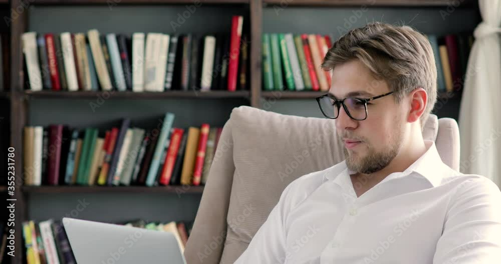 Portrait of man in glasses is working on computer on bookshelves background. University, College professor, businessman, office worker, freelancer is typing on laptop sitting on chair in library. Portrait of man in glasses is working on computer on bookshelves background. University, College professor, businessman, office worker, freelancer is typing on laptop sitting on chair in library.