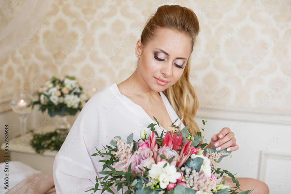Pretty young Bride. Blonde-haired woman with wedding hair-style with a long tail. Boudoir morning of the bride. Looking on her bouquet Pretty young Bride. Blonde-haired woman with wedding hair-style with a long tail. Boudoir morning of the bride. Looking on her bouquet
