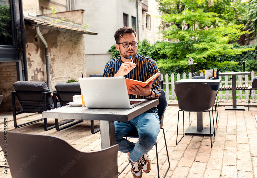 Portrait of man in casual wear sitting in cafe working using laptop and notebook.