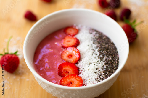 Healthy vegan trendy breakfast: magenta pink berry smoothie bowl made with frozen bananas, raspberry, strawberry and coconut milk. Topped with coconut flakes chia seeds and sliced strawberry. 