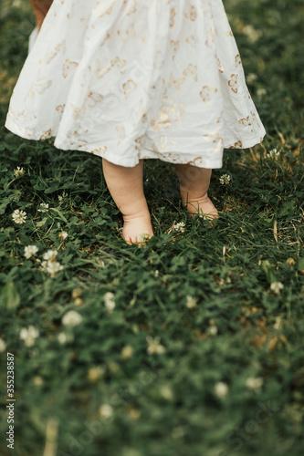 Cropped photo: barefoot baby girl in white dress standing on the green grass in park at summertime. The first steps in close-up. Healthy baby feet. First step. 