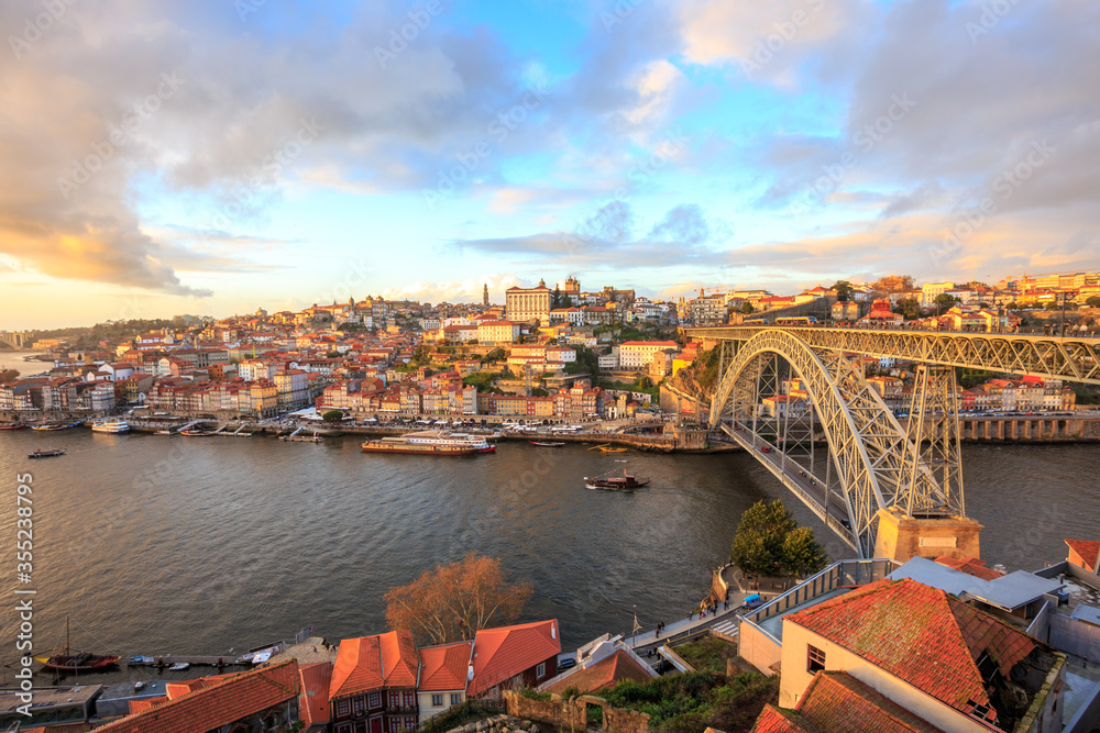 Afternoon in Porto Portugal: Douro river and Ribeira district featuring Ponte Luis I bridge and Episcopal palace on the top of the hill at sunset, under orange and blue clouds in the sky.