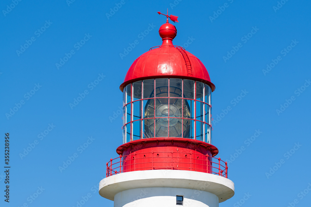 The top of a vintage lighthouse tower with a round red metal roof.  In the center of the lighthouse is a vintage lamp made of multiple pieces of glass. On top of the tower is a red metal arrow.