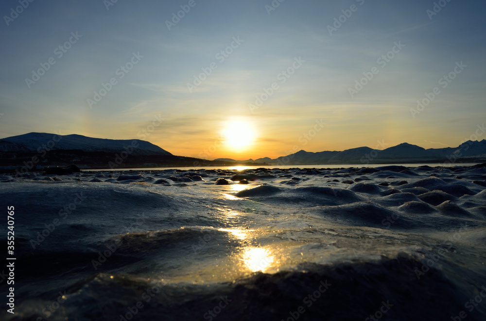 golden sunrise over blue fjord and snowy mountain with reflection on thick frozen sea shore ice