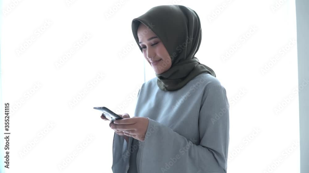 Beautiful hijab woman is typing a message on her phone. while standing near her window Beautiful hijab woman is typing a message on her phone. while standing near her window