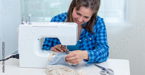A beautiful cheerful woman seamstress in a blue checked shirt, in her workshop, on a sewing machine, sewing a blue bra with beige inserts.