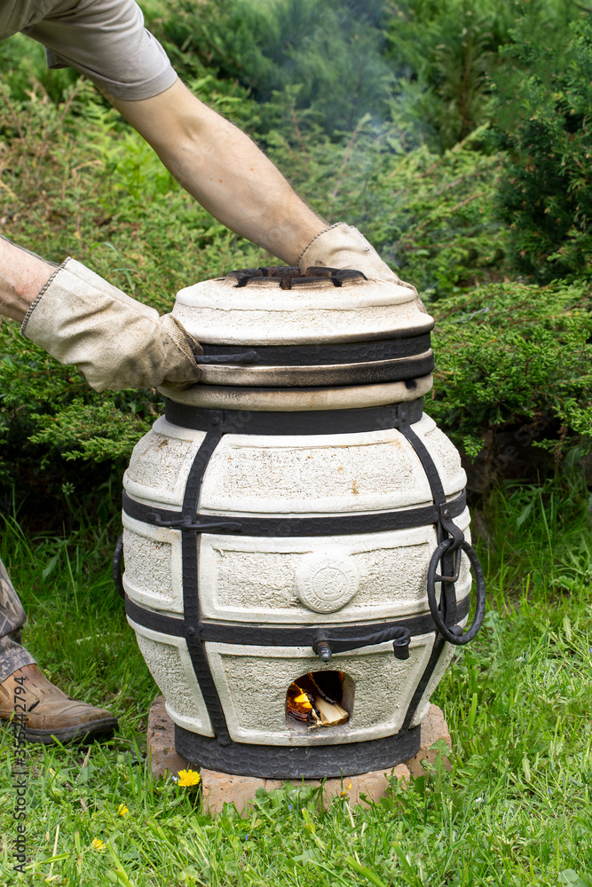 Burning tandoor stands on the grass.A man closes a lid on a smoking tandoor. Burning tandoor stands on the grass.A man closes a lid on a smoking tandoor.