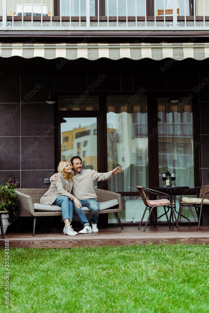 Obraz premium Mature man pointing with hand and embracing smiling wife on wicker sofa on terrace