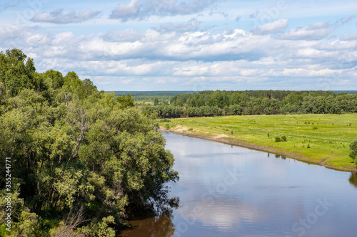 A small river flows around the forest, with the reflection of the trees in it against the blue sky. Nature of the Siberian expanses.