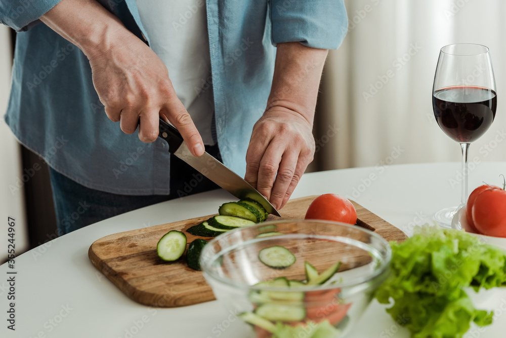 Cropped view of man cutting cucumber while cooking salad near glass of wine in kitchen