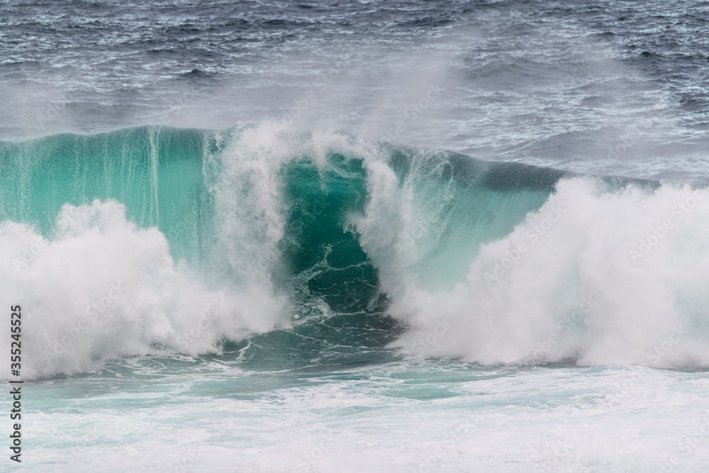 A large green teal coloured wave rolling in on a beach. The wave has white splashing foam from the crashing as the ocean meets the beach. The rip curl of the wave is a barrel wave rolling in onshore. 