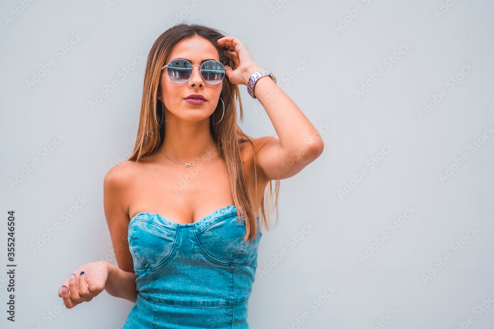 Lifestyle session, a young caucasian brunette in a blue denim dress and sunglasses on a plain gray background Lifestyle session, a young caucasian brunette in a blue denim dress and sunglasses on a plain gray background