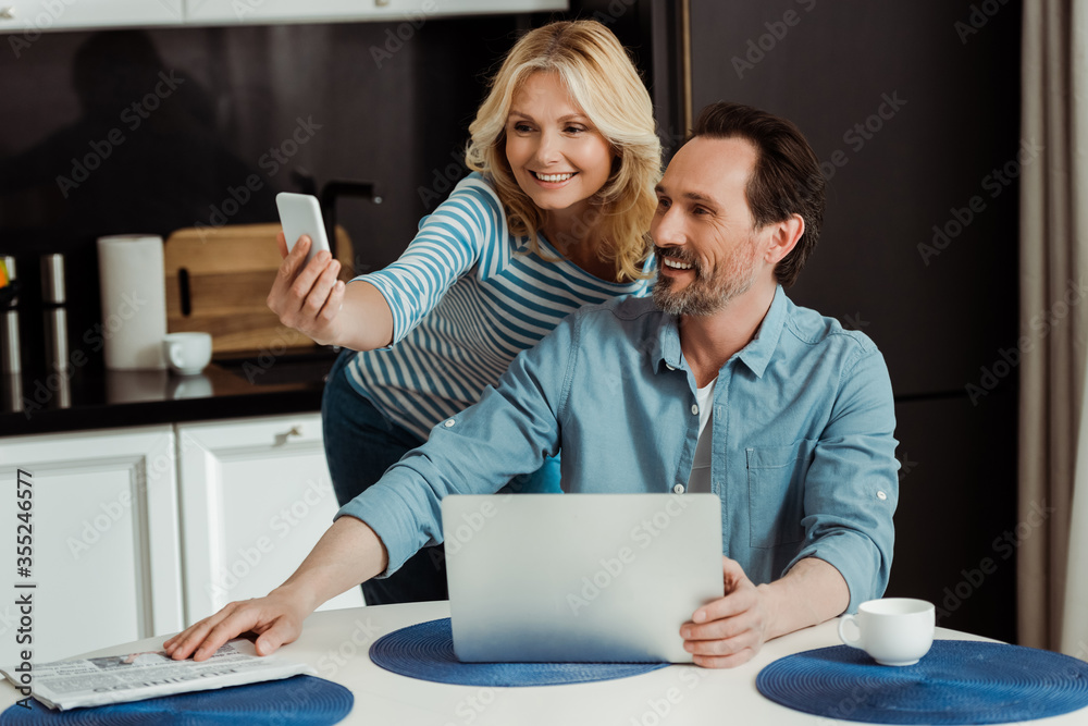 Smiling woman taking selfie with smartphone near husband using laptop in kitchen Smiling woman taking selfie with smartphone near husband using laptop in kitchen