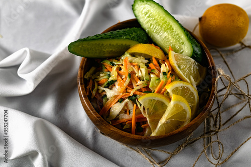 Healthy salad of fresh vegetables in a deep wooden plate. A dish with white cabbage, carrots and fragrant cucumbers, decorated with lemon slices and herbs. Interesting presentation of healthy eating