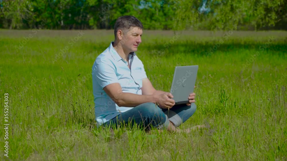 Man Works behind a Laptop Sitting on the Grass At The Sunny Day. Freelancer Remote Work Concept. 4K Man Works behind a Laptop Sitting on the Grass At The Sunny Day. Freelancer Remote Work Concept. 4K