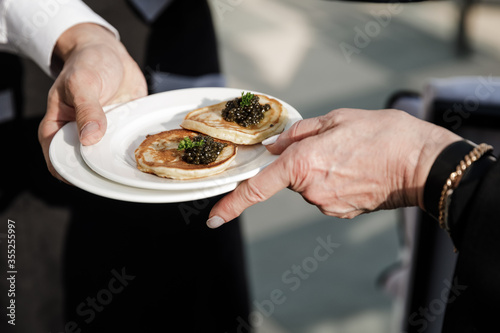 The waiter passes a plate of pancakes and black caviar to the guest. 