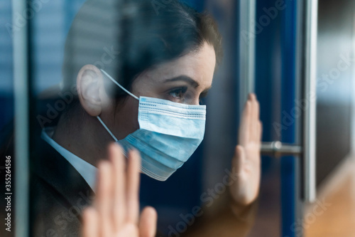A sad quarantined woman in a business suit leaned against the window. Caucasian girl in a mask on isolation put her hands on a glass door. Maintaining social distance to prevent coronavirus.