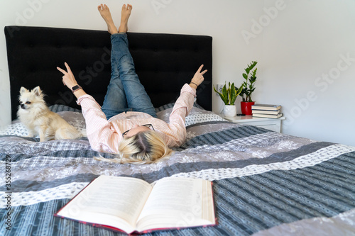 Foto Blonde hair woman lying legs up on cozy bed with dog, holding open book and reading