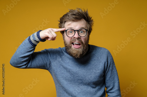 Irritated shaggy man with glasses holds a finger near his temple, showing interlocutor that he is an idiot. photo