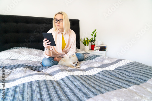 Foto Blonde hair woman sitting on cozy bed with dog, holding e-book and reading