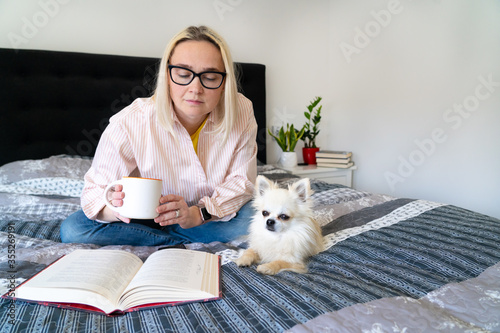 Bild auf Leinwand Blonde hair woman sitting on cozy bed with dog, holding open book and reading