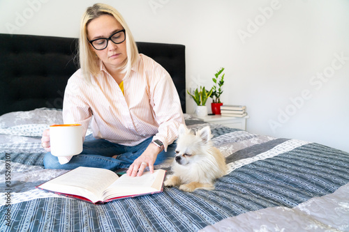 Foto Blonde hair woman sitting on cozy bed with dog, holding open book and reading