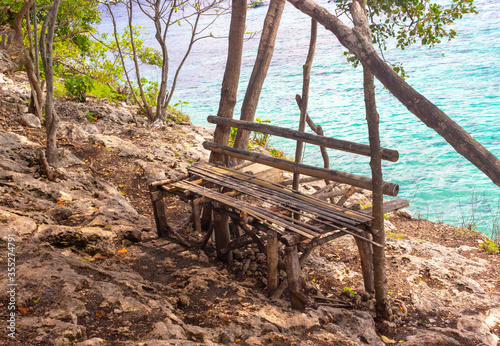 Secluded bench for relaxing on the rocky shore of the azure sea under the shadow of tropical trees