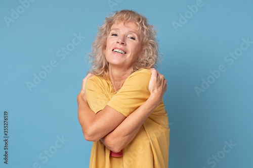 Relaxed happy mature woman hugging herself, having pleased joyful facial expression. Self love concept. Studio shot on blue wall.
